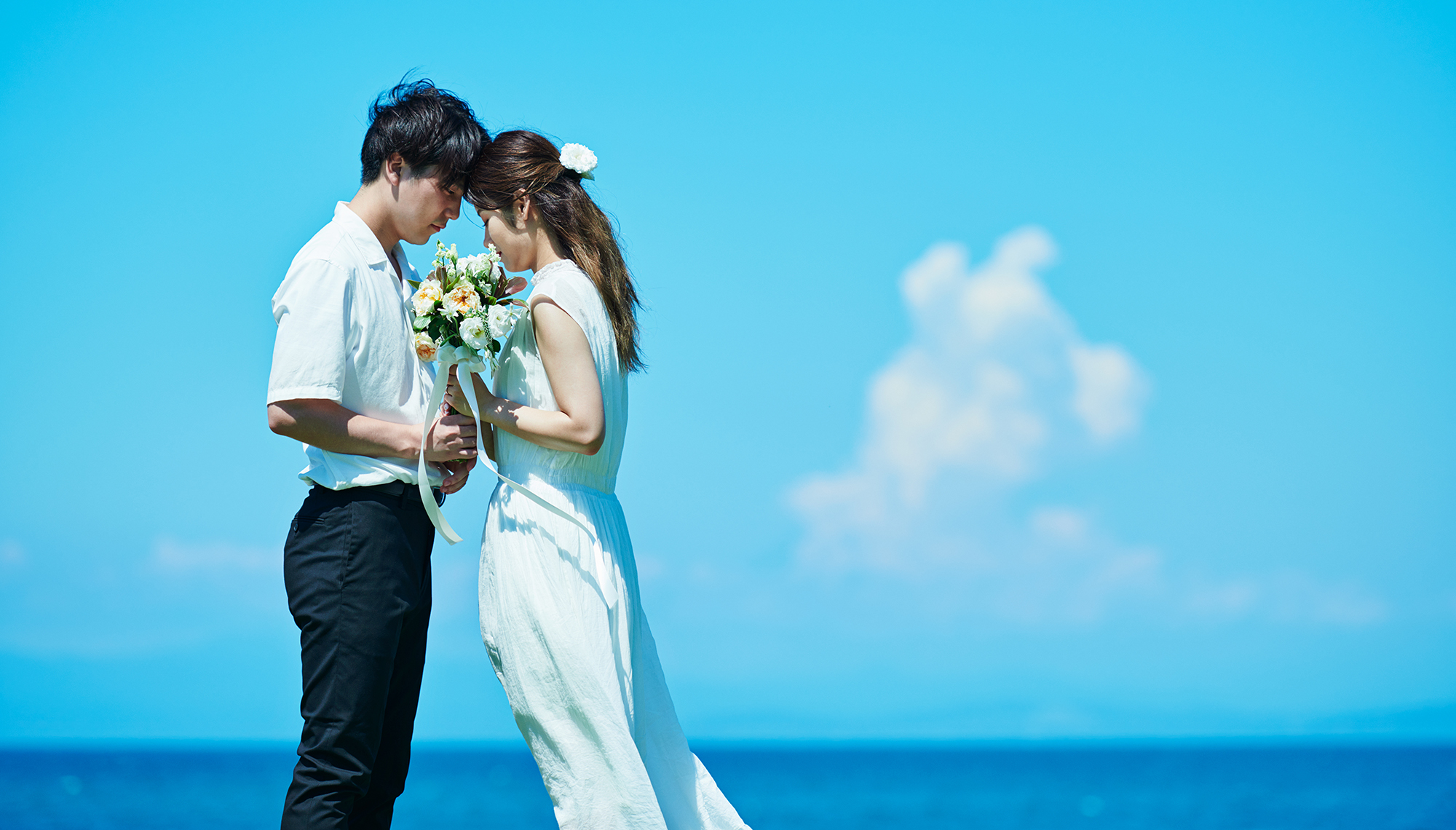 青い空が広がる海辺に、ブーケを持った恋人同士が額を付けあって向き合っている謝意🅂ン